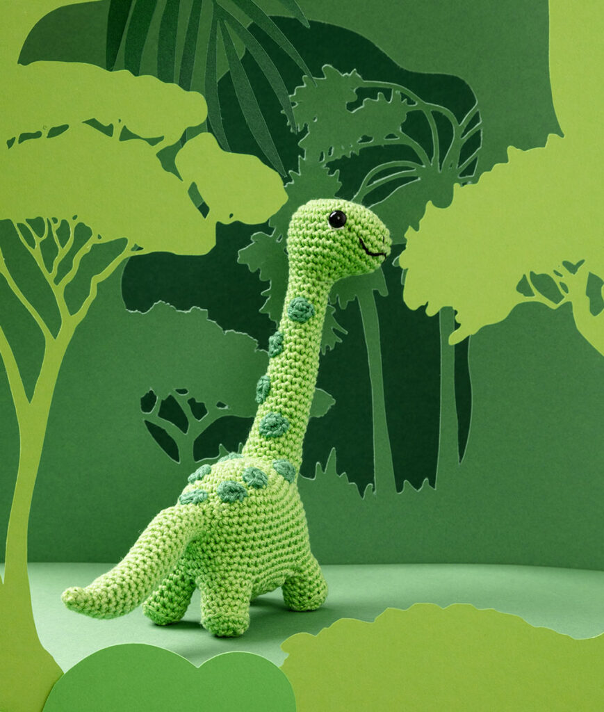 Produktfoto eines grünen Dinosauriers in grüner Paper-Art Kulisse mit verschiedenen Ebenen, die eine Landschaft der Urzeit ergeben