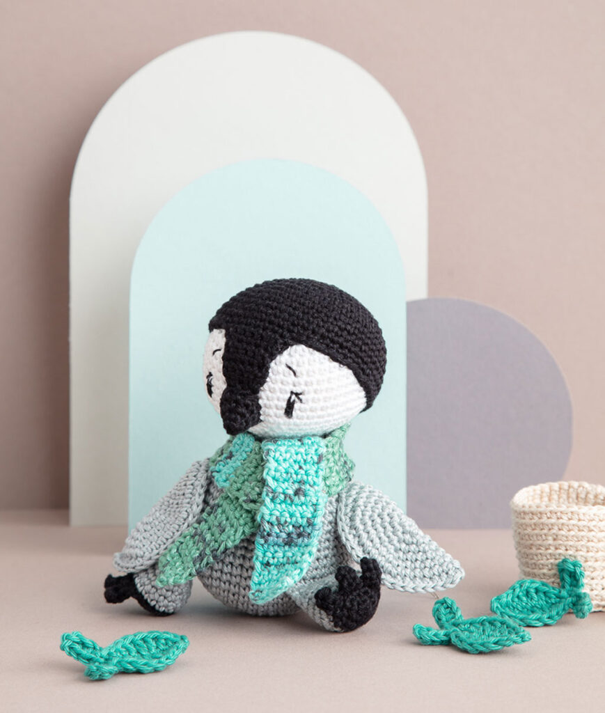 inszeniertes Produktfoto einer bekleideten Pinguin-Häkelpuppe vor einem Paper-Art Hintergrund mit verschiedenfarbigen runden, organischen Formen für ein Häkelbuch
