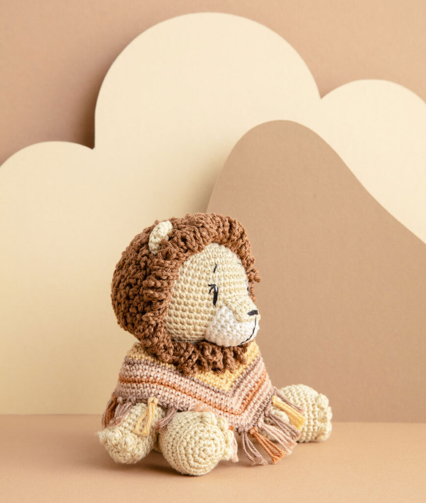 inszeniertes Produktfoto einer bekleideten Löwen-Häkelpuppe vor einem Paper-Art Hintergrund mit verschiedenfarbigen runden, organischen Formen für ein Häkelbuch