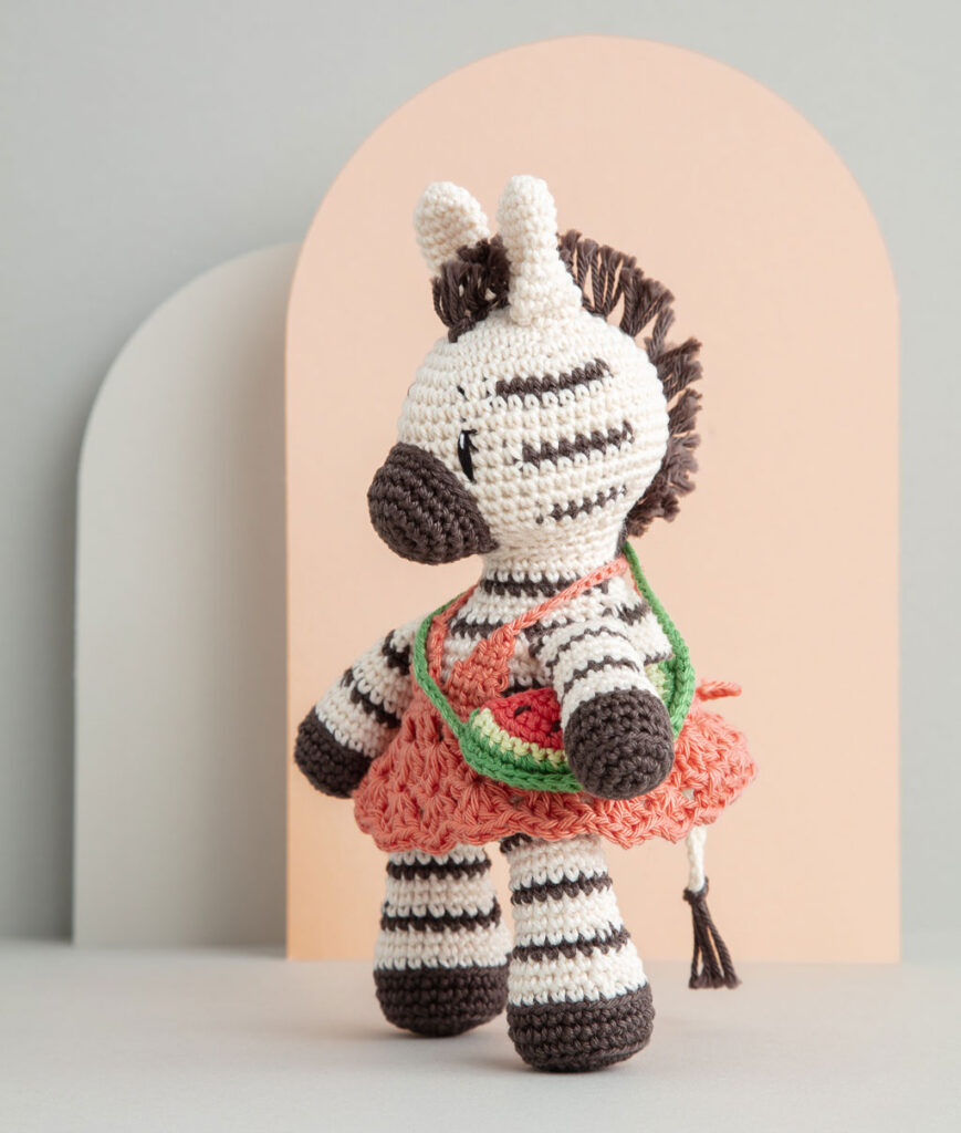 inszeniertes Produktfoto einer bekleideten Zebra-Häkelpuppe vor einem Paper-Art Hintergrund mit verschiedenfarbigen runden, organischen Formen für ein Häkelbuch