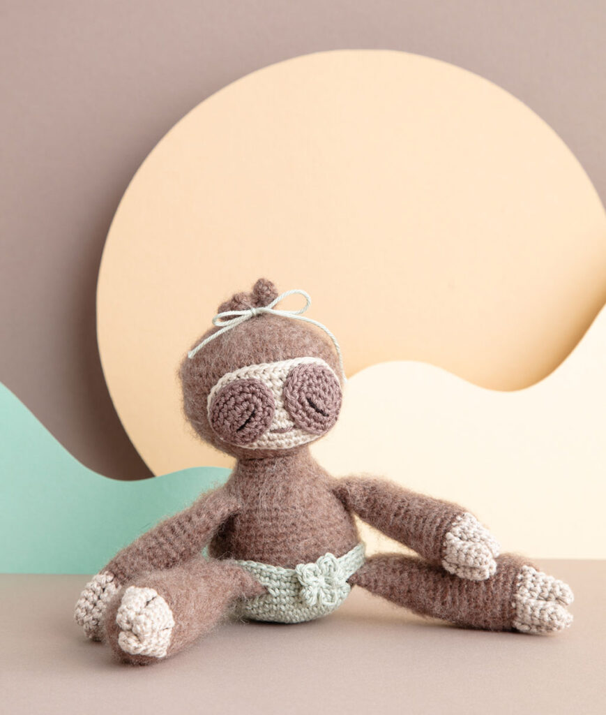inszeniertes Produktfoto einer bekleideten Faultier-Häkelpuppe vor einem Paper-Art Hintergrund mit verschiedenfarbigen runden, organischen Formen für ein Häkelbuch
