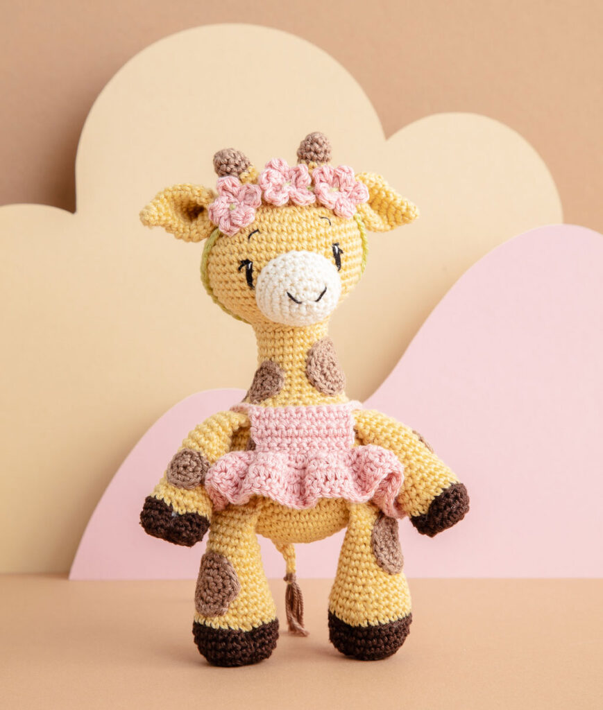 inszeniertes Produktfoto einer bekleideten Giraffe-Häkelpuppe vor einem Paper-Art Hintergrund mit verschiedenfarbigen runden, organischen Formen für ein Häkelbuch