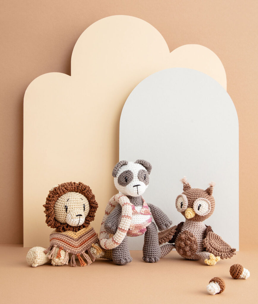 inszeniertes Produktfoto mit drei bekleideten Häkeltieren - ein Löwe, ein Pandabär und eine Eule - vor einem Paper-Art Hintergrund mit verschiedenfarbigen runden, organischen Formen für das Coverfoto eines Häkelbuch