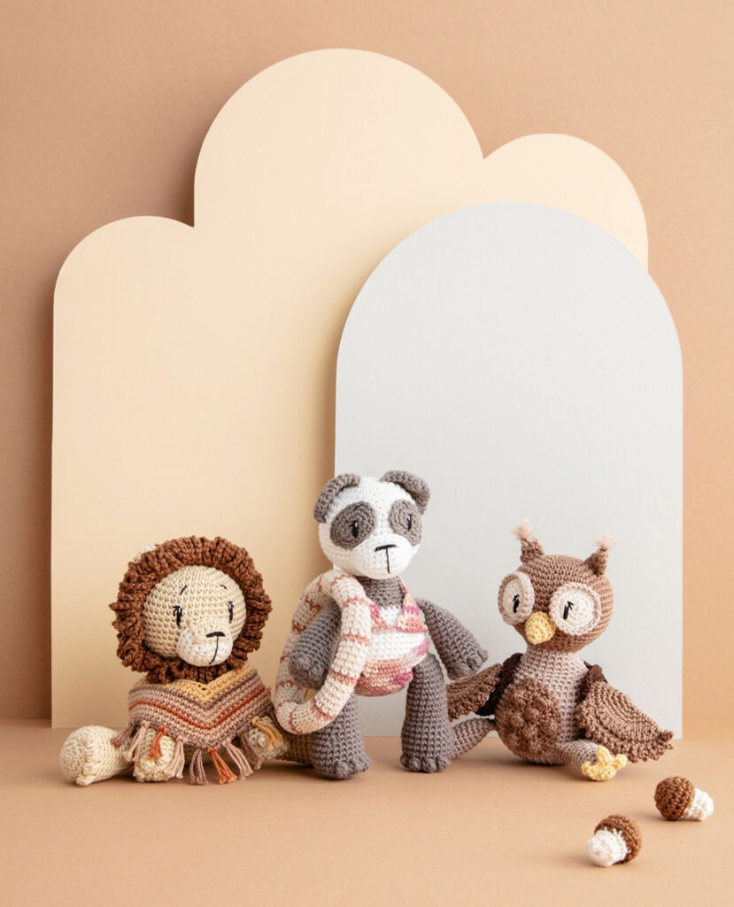 inszeniertes Coverfoto mit drei bekleideten Häkeltieren - Löwe, Pandabär und Eule - vor einem Paper-Art Hintergrund mit verschiedenfarbigen runden, organischen Formen für ein Häkelbuch