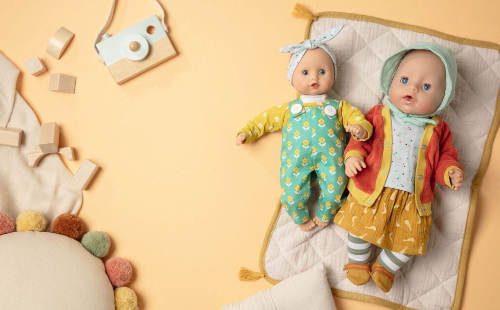 Produktfoto als Flatlay für Puppenkleidung mit zwei bekleideten Puppen auf gelbem Untergrund mit Kissen, Decke und Holzbausteinen