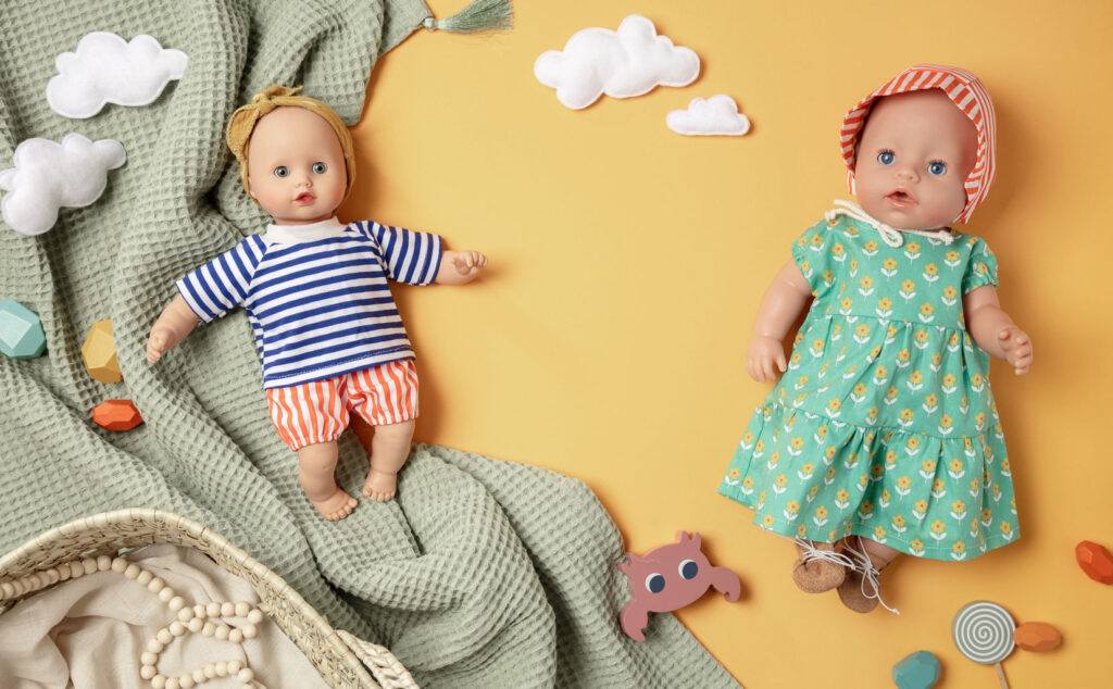 sommerliches Produktfoto als Flatlay für Puppenkleidung mit zwei bekleideten Puppen auf gelbem Untergrund mit Decke, Körbchen, Holzbausteinen und Filzwolken