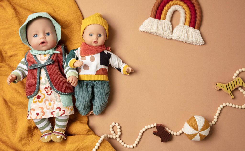 herbstliches Produktfoto als Flatlay für Puppenkleidung mit zwei bekleideten Puppen auf braunem Untergrund mit Decke, Holzkugelkette und Deko-Regenbogen