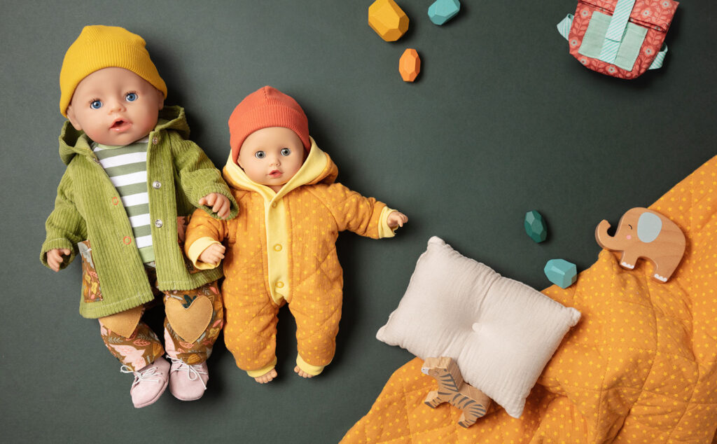 winterliches Produktfoto als Flatlay für Puppenkleidung mit zwei bekleideten Puppen auf grünem Untergrund mit Kissen, Decke und Holzbausteinen