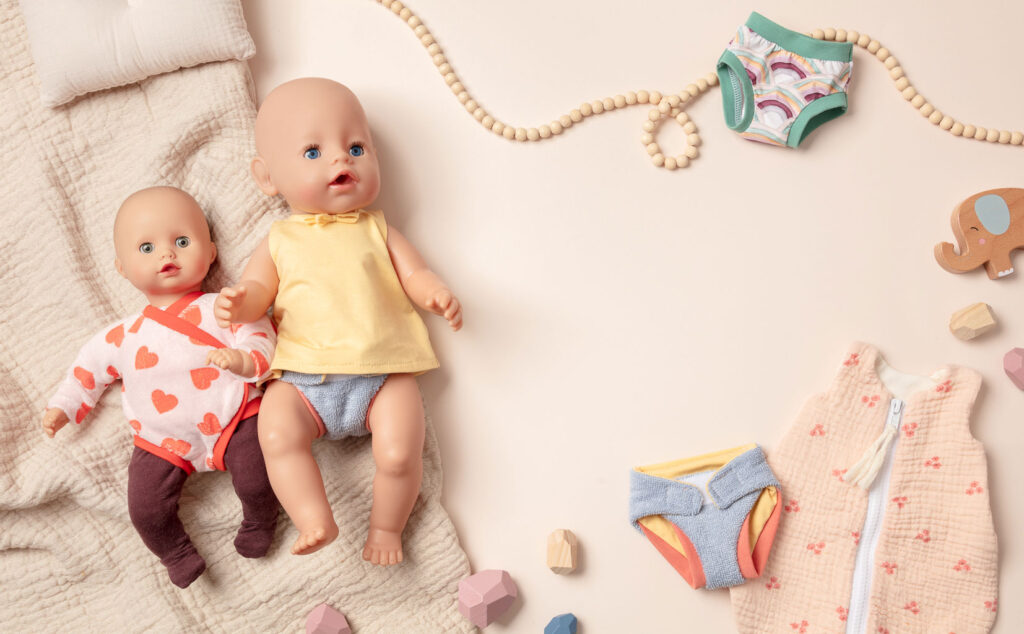 Produktfoto als Flatlay für Puppenkleidung mit zwei bekleideten Puppen auf hellem Untergrund mit Kissen, Wäsche und Holzdeko