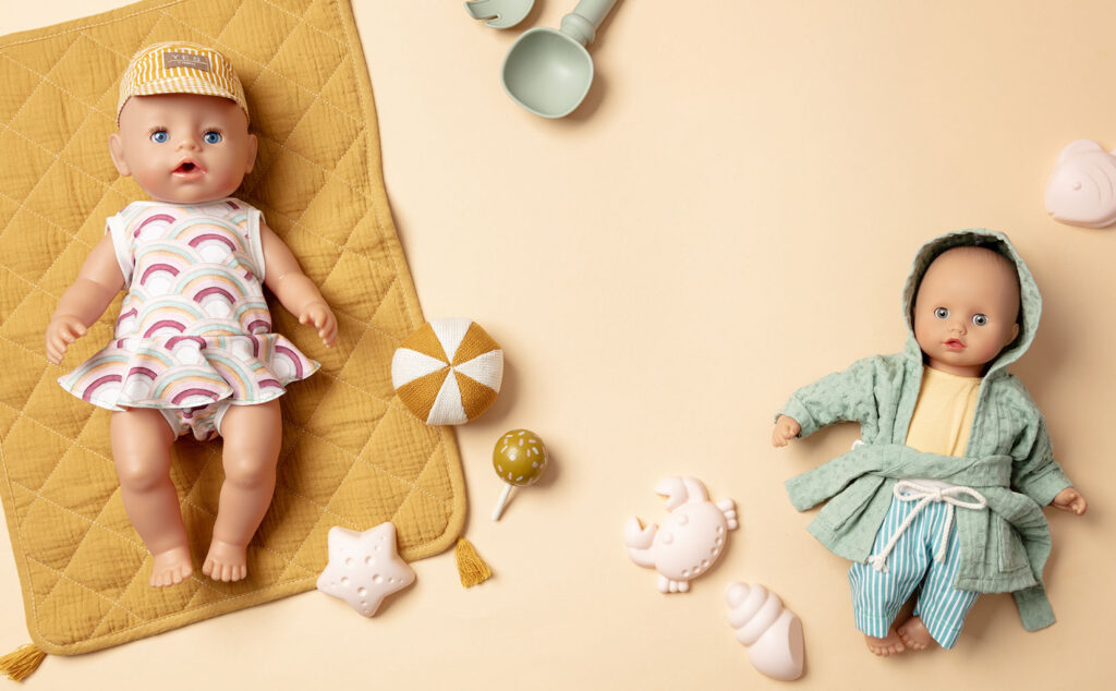 sommerliches Produktfoto als Flatlay für Puppenkleidung mit zwei bekleideten Puppen auf gelbem Untergrund mit Decke und Sandspielzeug