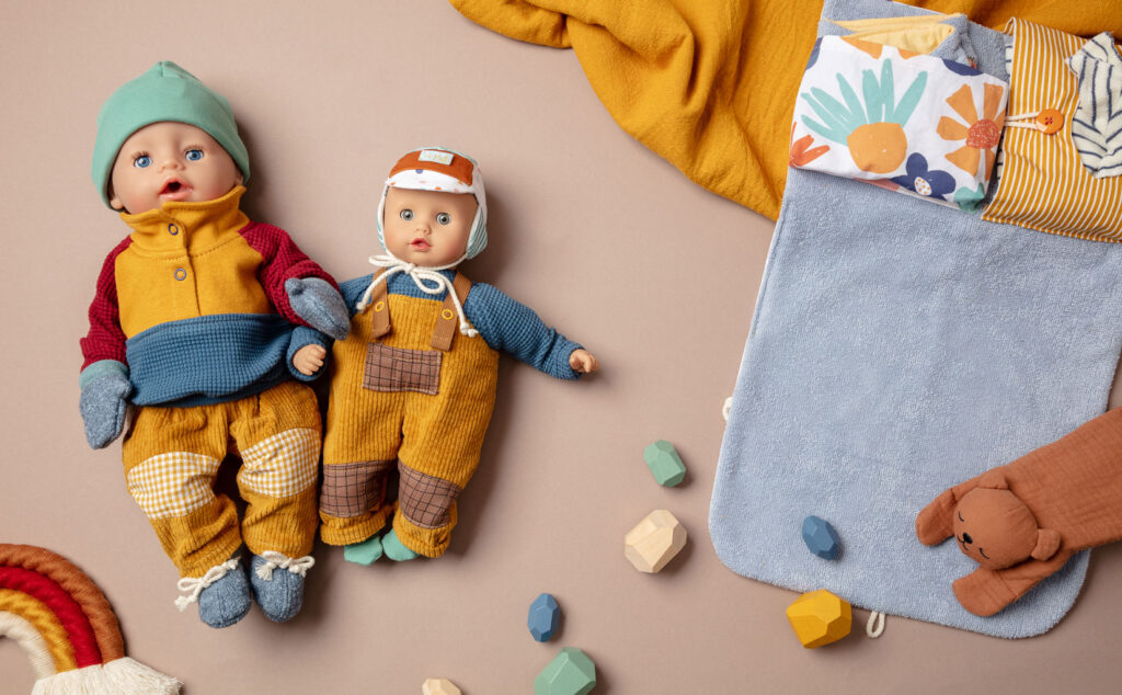 herbstliches Produktfoto als Flatlay für Puppenkleidung mit zwei bekleideten Puppen auf braunem Untergrund mit Wickeltasche, Decke und Holzbausteinen