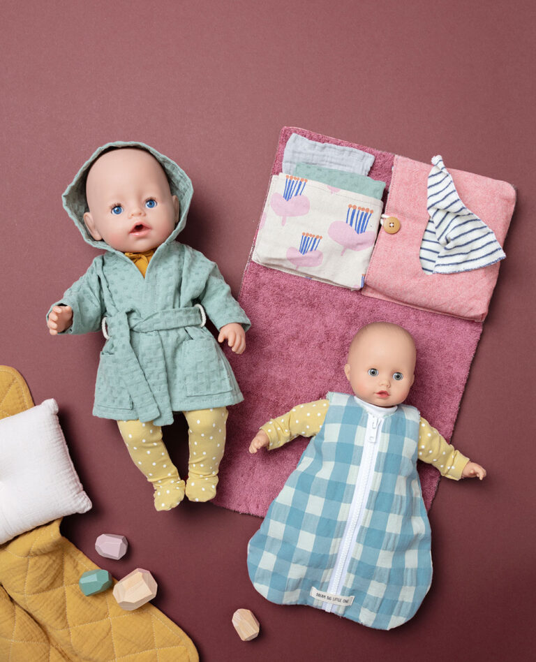 Produktfoto als Flatlay für Puppenkleidung mit zwei bekleideten Puppen auf beerenfarbigen Untergrund mit Kissen, Decke und Wickelsachen