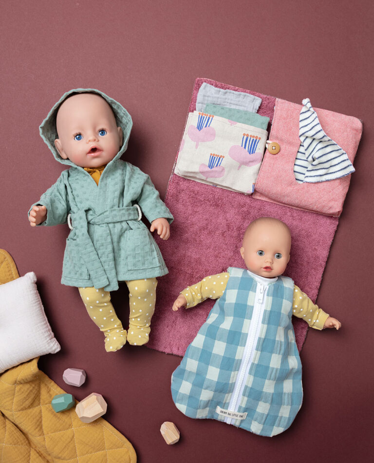 Titelfoto für Puppenkleidung mit zwei bekleideten Puppen auf beerenfarbigen Untergrund mit Kissen, Decke und Wickelsachen