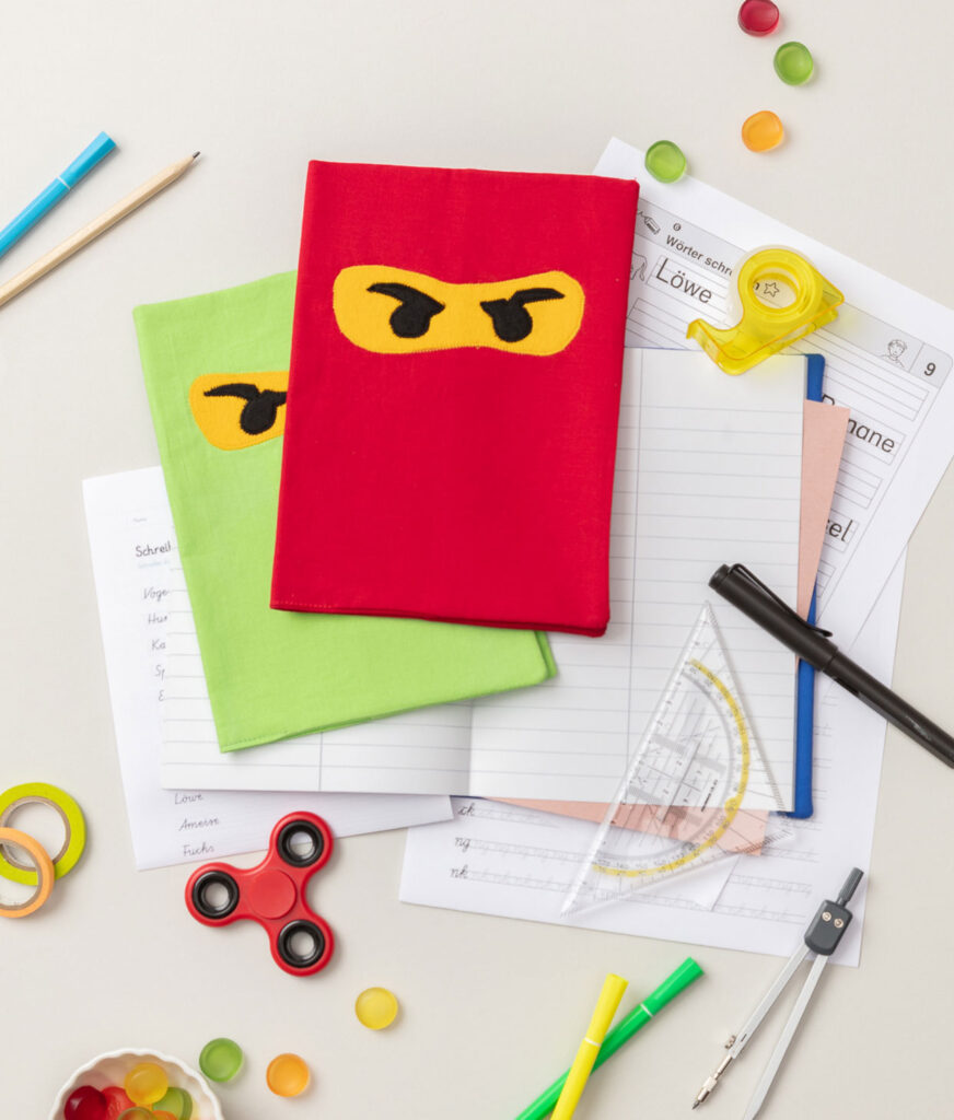 Produktfoto zweier genähter Hefthüllen mit Ninja Augen auf grauem Hintergrund dekoriert mit Schulheften, Stiften und Schulmaterialien für ein Kinder Nähbuch