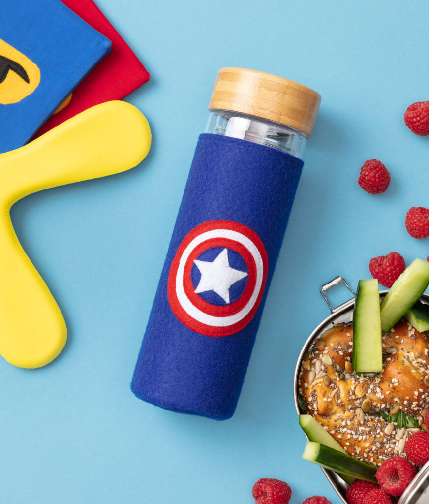 Produktfoto eines genähten Flaschenschutzes mit Avenger-Symbol auf blauem Hintergrund dekoriert mit Brotdose, Heften und Frisbee für ein Kinder Nähbuch