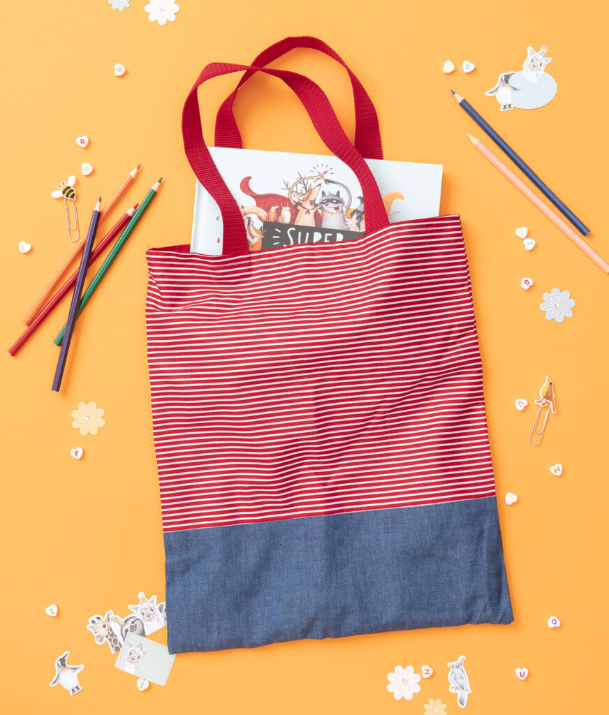 Produktfoto eines genähten Beutels in Conni-Farben auf gelbem Hintergrund dekoriert mit Freundebuch, Stiften und Stickern für ein Kinder Nähbuch
