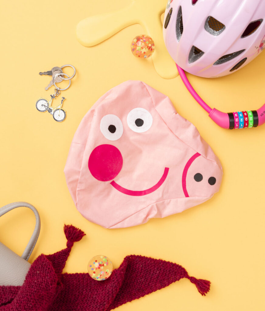 Produktfoto eines genähten Sattelüberziehers als Peppa Wutz Figur auf gelben Hintergrund dekoriert mit Kinderhelm, Fahrradschloss, Schlüssel und bunten Bällen für ein Kinder Nähbuch