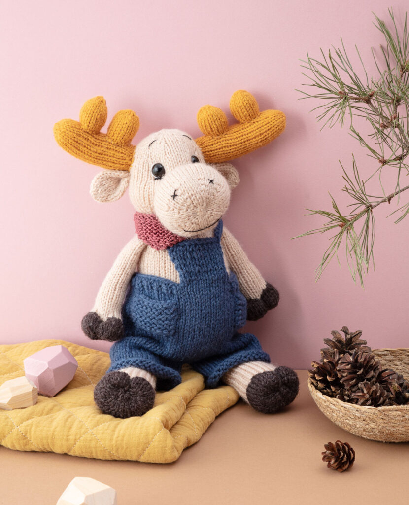inszeniertes Produktfoto eines verniedlicht dargestellten, bekleideten Elches vor rosanem Hintergrund mit Tannenzweig, einer Schale Zapfen, Holzsteinen und einer Decke für ein Strickbuch