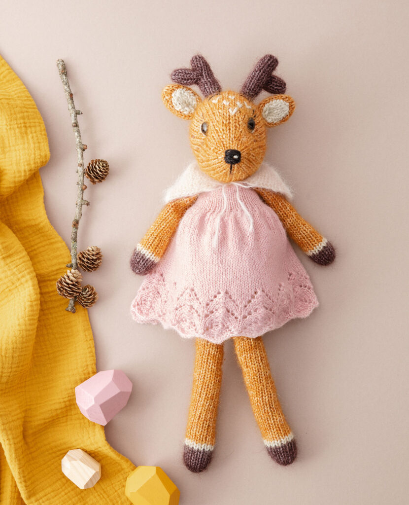 inszeniertes Produktfoto eines verniedlicht dargestellten, bekleideten Rehs als Flatlay auf hellgrauem Hintergrund mit Lärchenzweig, Holzdeko und gelber Decke für ein Strickbuch