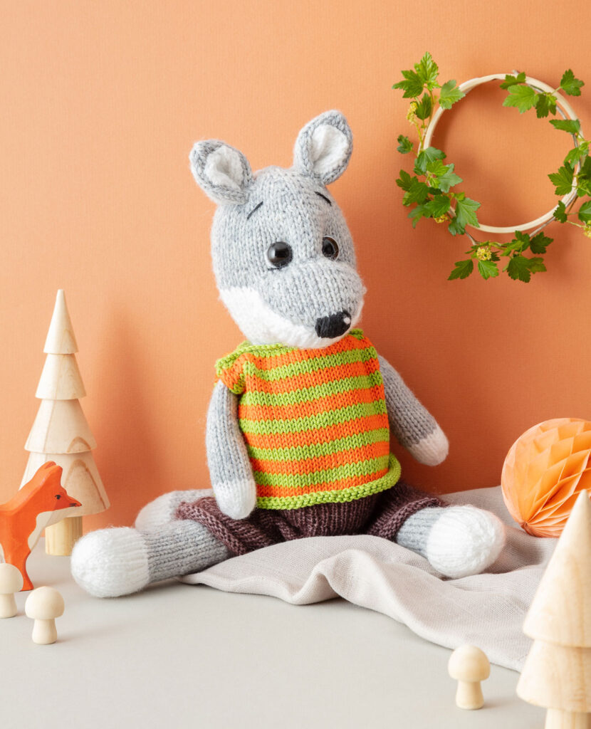 inszeniertes Produktfoto eines verniedlicht dargestellten, bekleideten Fuchses vor orangem Hintergrund mit Blätterkranz, passender Holzdeko und Musselindecke für ein Strickbuch
