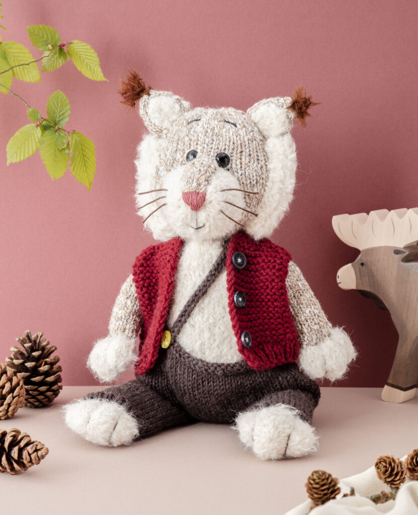 inszeniertes Produktfoto eines verniedlicht dargestellten, bekleideten Luchses vor weinrotem Hintergrund mit Zweigen, Elchdeko und Tannenzapfen für ein Strickbuch