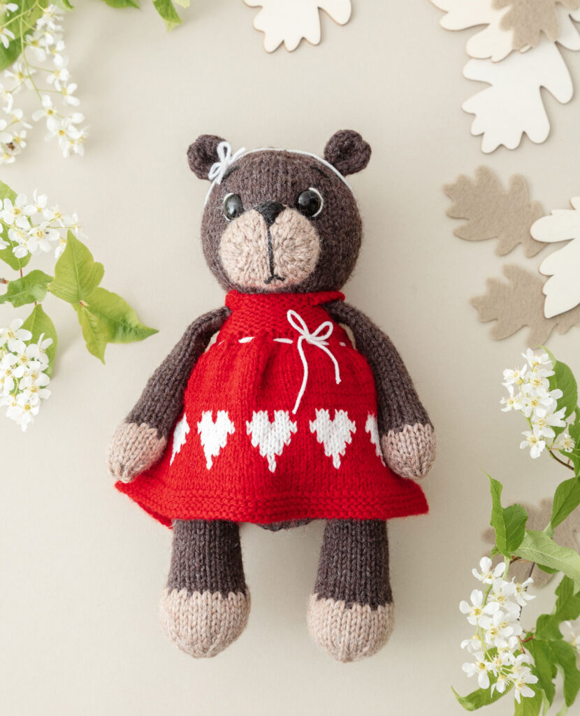 inszeniertes Produktfoto einer verniedlicht dargestellten, bekleideten Bärendame als Flatlay auf hellgrauem Hintergrund mit Blüten, Grün und Blätterdekoration für ein Strickbuch