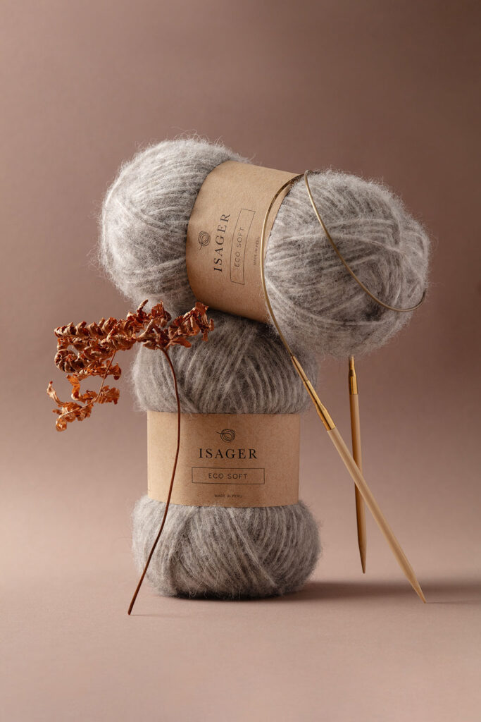 Stimmungsvolles Produktfoto von zwei Knäuel Eco Soft Wolle von Isager mit einer Rundstricknadel, dekoriert mit einem vertrockneten Zweig Farn vor einem dunkelbraunem Hintergrund