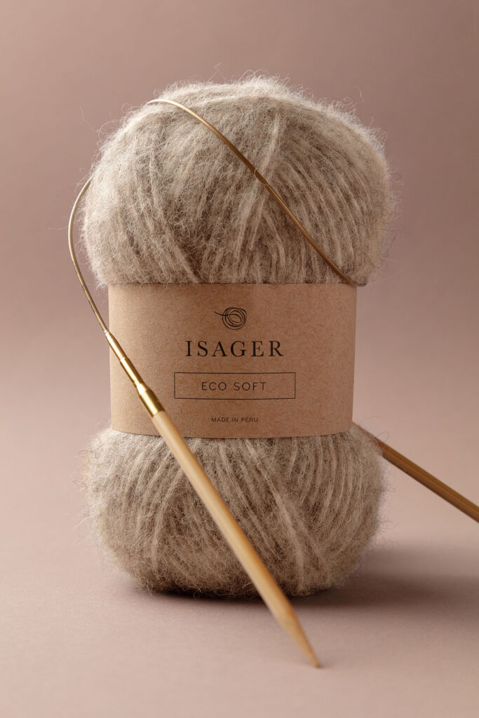 Stimmungsvolles Produktfoto von einem Knäuel Eco Soft Wolle von Isager, dekoriert mit einer Rundstricknadel, vor einem braunem Hintergrund