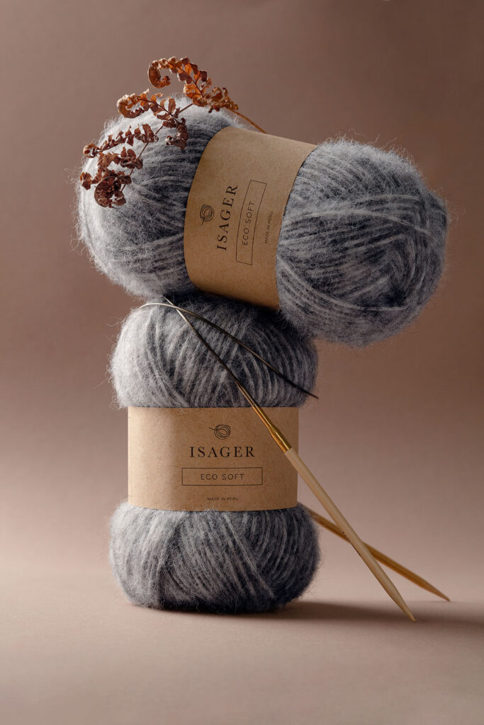 Stimmungsvolles Produktfoto von zwei Knäuel Eco Soft Wolle von Isager mit einer Rundstricknadel, dekoriert mit einem vertrockneten Zweig Farn vor einem dunkelbraunem Hintergrund