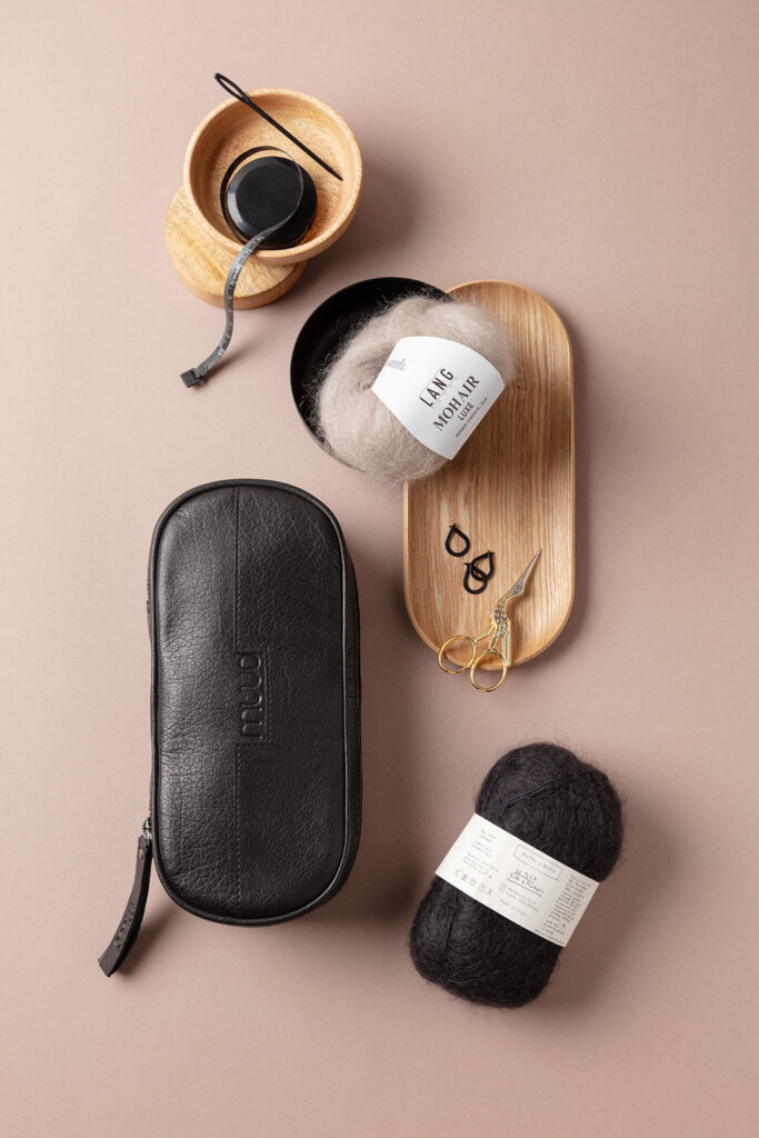 Arrangiertes Flatlay-Produktfoto auf braunem Untergrund, das zwei unterschiedliche Knäuel Mohair-Wolle zeigt, dekoriert mit einer schwarzen Zubehörtasche von Muud, einer Holzschale mit weiterem Strickzubehör