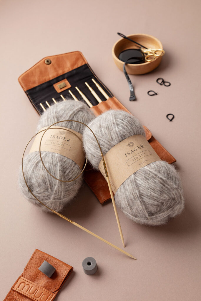 Arrangiertes Produktfoto von zwei Knäuel Eco Soft Wolle von Isager , dekoriert mit einer Rundstricknadel, Zubehörtaschen von muud und einer Holzschale mit weiterem Strickzubehör