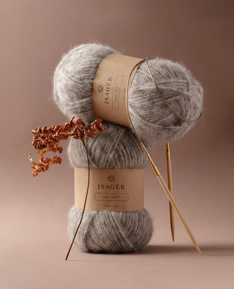 Stimmungsvolles Produktfoto von zwei Knäuel Eco Soft Wolle von Isager mit einer Rundstricknadel, dekoriert mit einem vertrockneten Zweig Farn vor einem dunkelbraunem Hintergrund