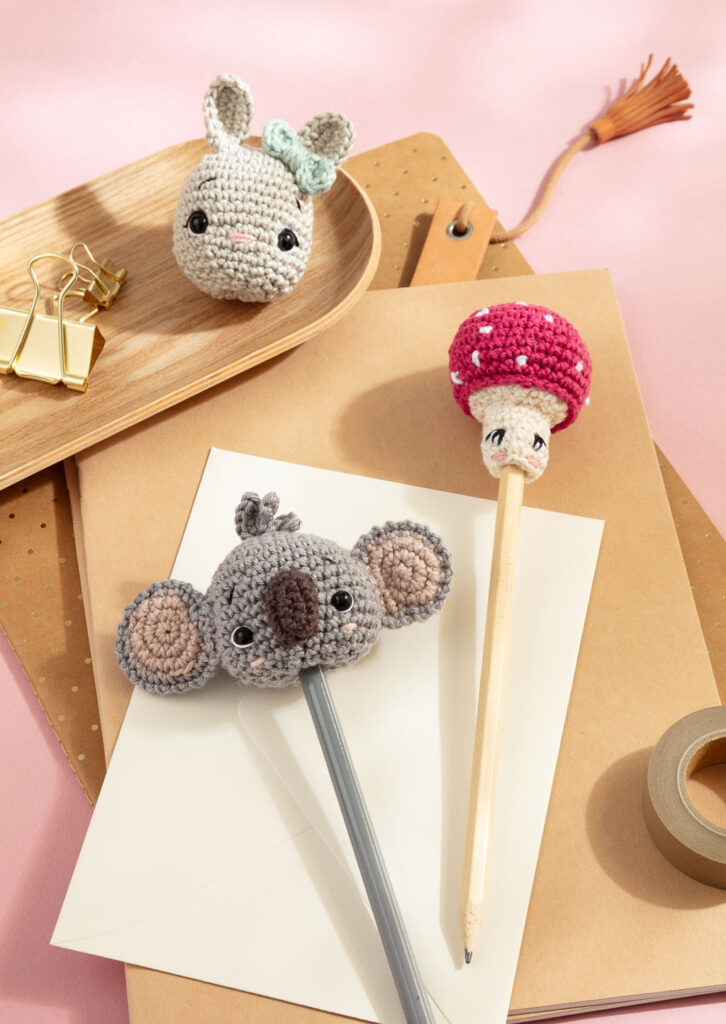Produktfoto für ein Häkelbuch, das drei gehäkelte Amigurumi-Stiftaufsätze auf dem Schreibtisch zeigt