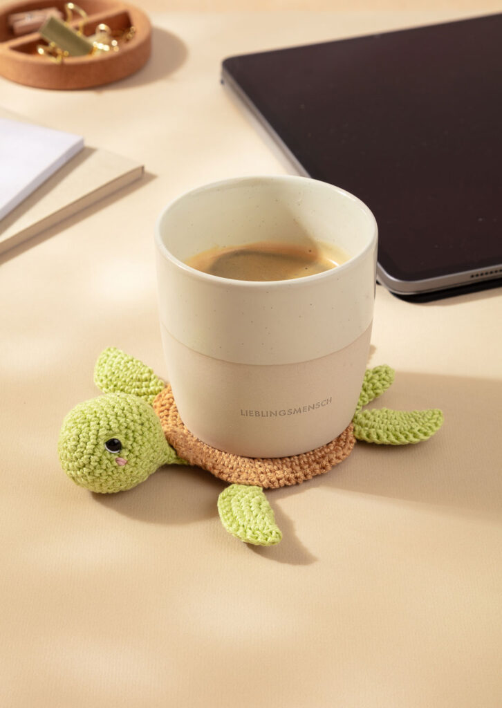 Produktfoto für ein Häkelbuch, das einen gehäkelten Amigurumi-Tassenuntersetzer auf einem Schreibtisch zeigt