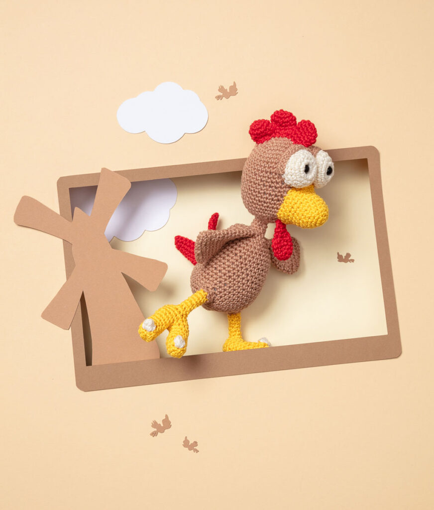Produktfoto einer Amigurumi-Figur, die das Moorhuhn aus dem Videospiel Moorhuhn darstellt, arrangiert in einer Kulisse aus einem Papier Cut-Out in Form eines Bildschirms, dekoriert mit weiteren spieltypischen Paper-Art-Elementen