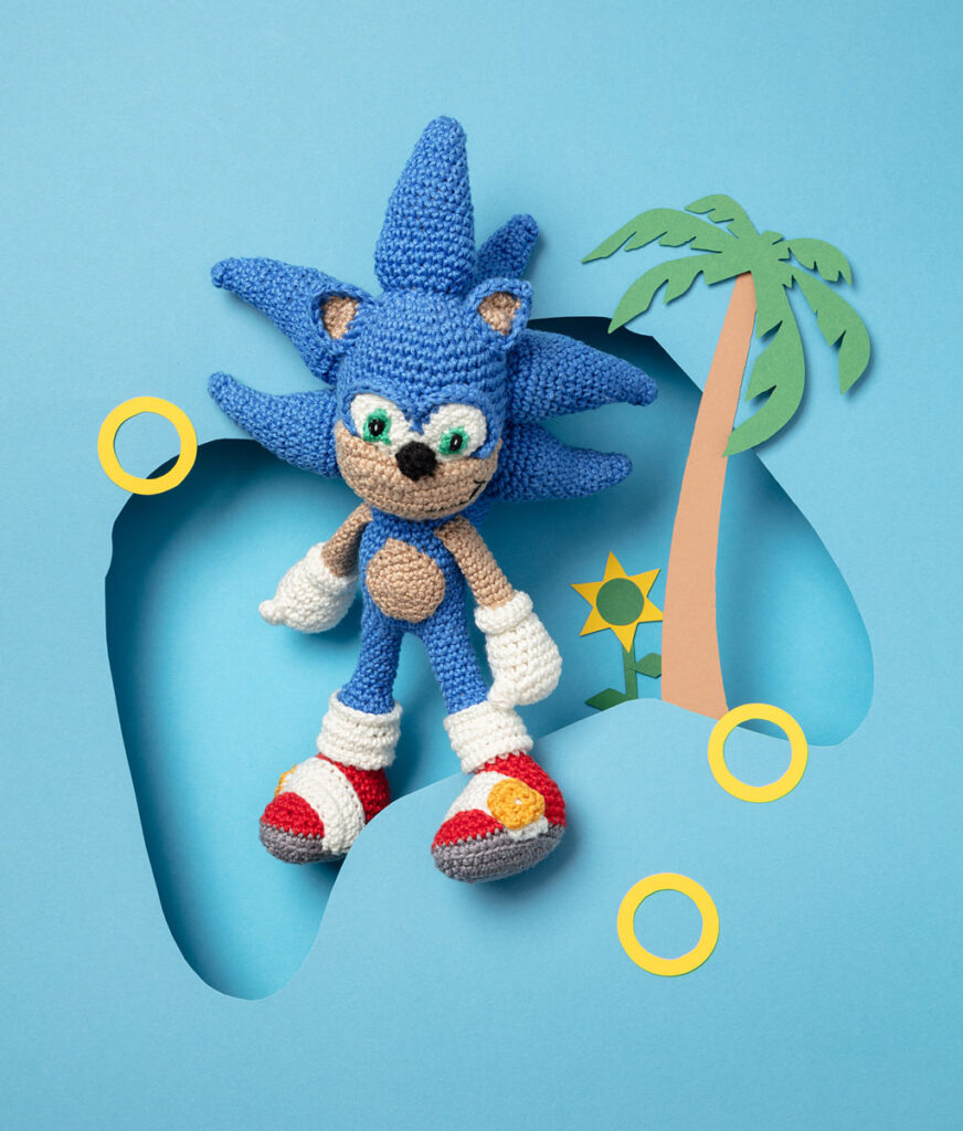 Produktfoto eines Amigurumis, das Sonic aus dem Videospiel Sonic the Hedgehog darstellt, arrangiert in einer Kulisse aus einem Papier Cut-Out in Form eines Controllers, dekoriert mit weiteren spieltypischen Paper-Art-Elementen