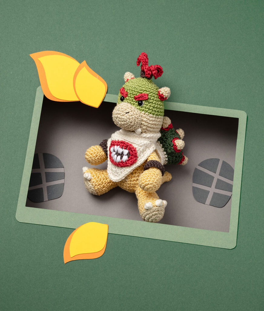 Produktfoto eines Amigurumis, das Baby Bowser aus den Videospielen Super Mario World darstellt, arrangiert in einer Kulisse aus einem Papier Cut-Out in Form eines Bildschirms, dekoriert mit weiteren spieltypischen Paper-Art-Elementen