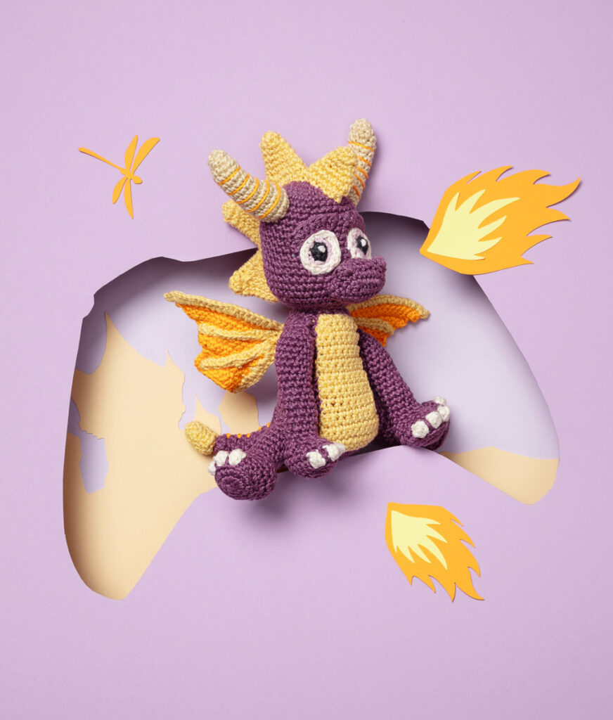 Produktfoto eines Amigurumis, das den Drachen aus dem Videospiel Spyro darstellt, arrangiert in einer Kulisse aus einem Papier Cut-Out in Form eines Controllers, dekoriert mit weiteren spieltypischen Paper-Art-Elementen