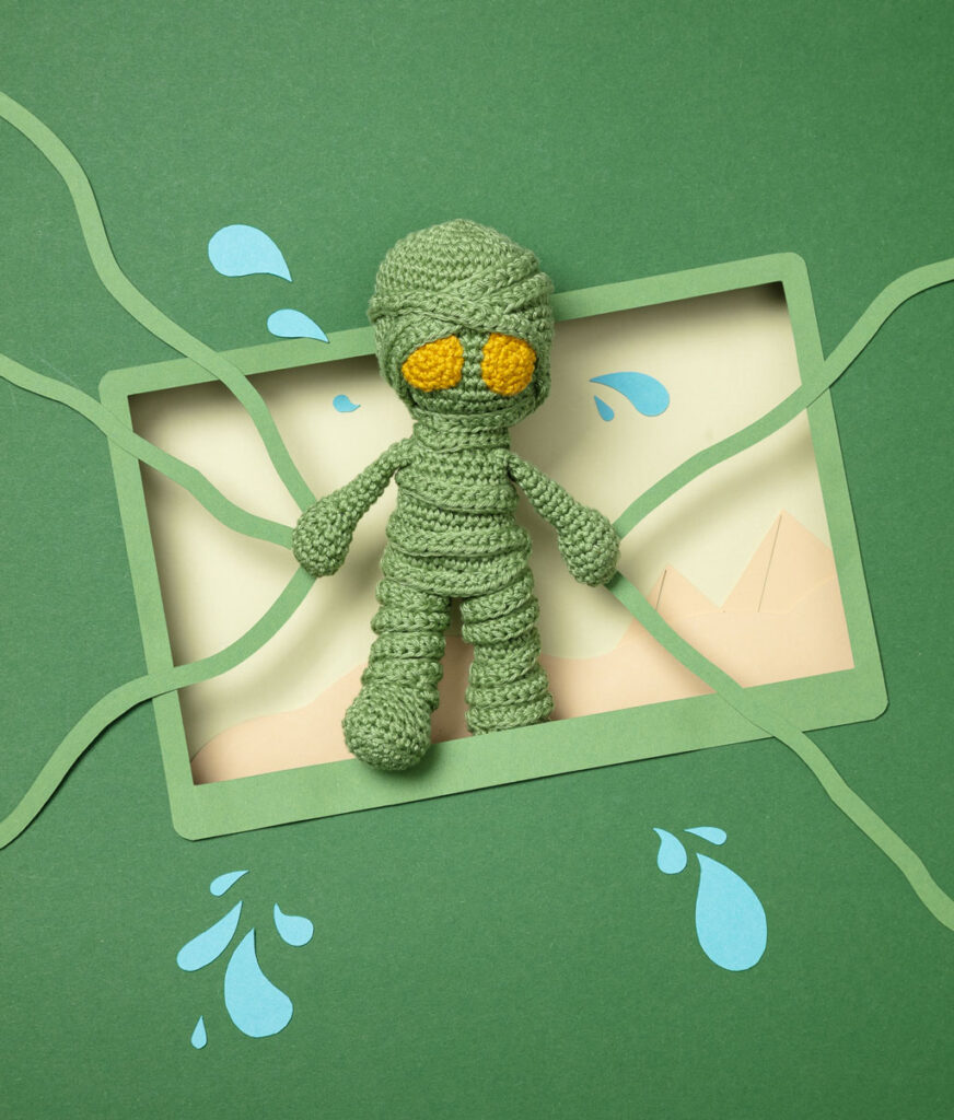 Produktfoto einer Amigurumi-Figur, die Amumu aus dem Videospiel League of Legends darstellt, arrangiert in einer Kulisse aus einem Papier Cut-Out in Form eines Bildschirms, dekoriert mit weiteren spieltypischen Paper-Art-Elementen