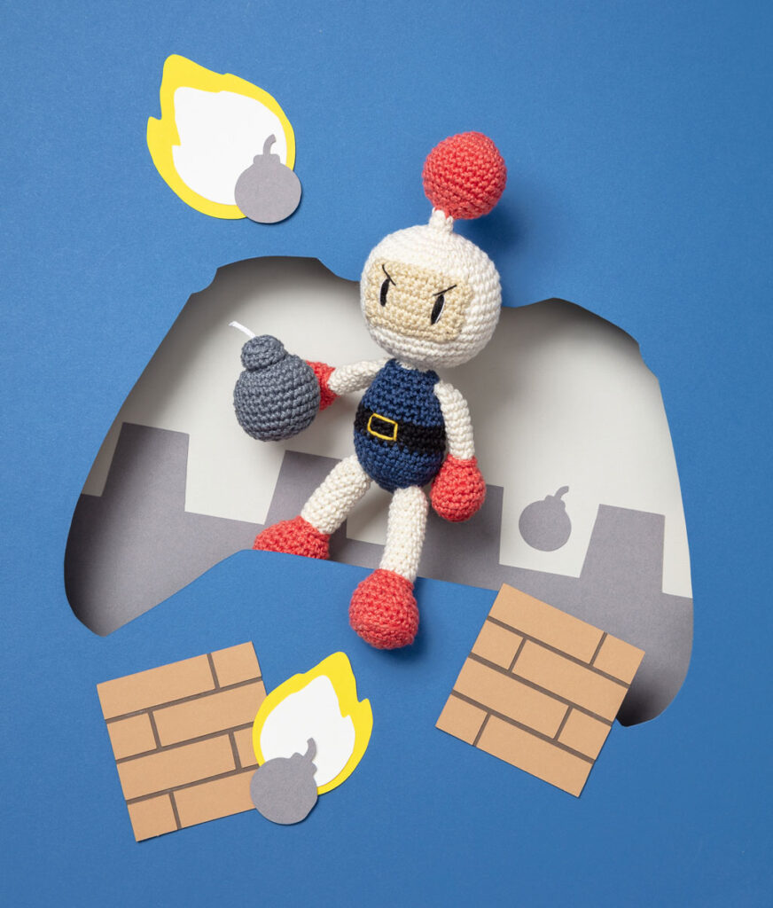 Produktfoto einer Amigurumi-Figur, die Bomberman aus dem Videospiel Bomberman darstellt, arrangiert in einer Kulisse aus einem Papier Cut-Out in Form eines Controllers, dekoriert mit weiteren spieltypischen Paper-Art-Elementen