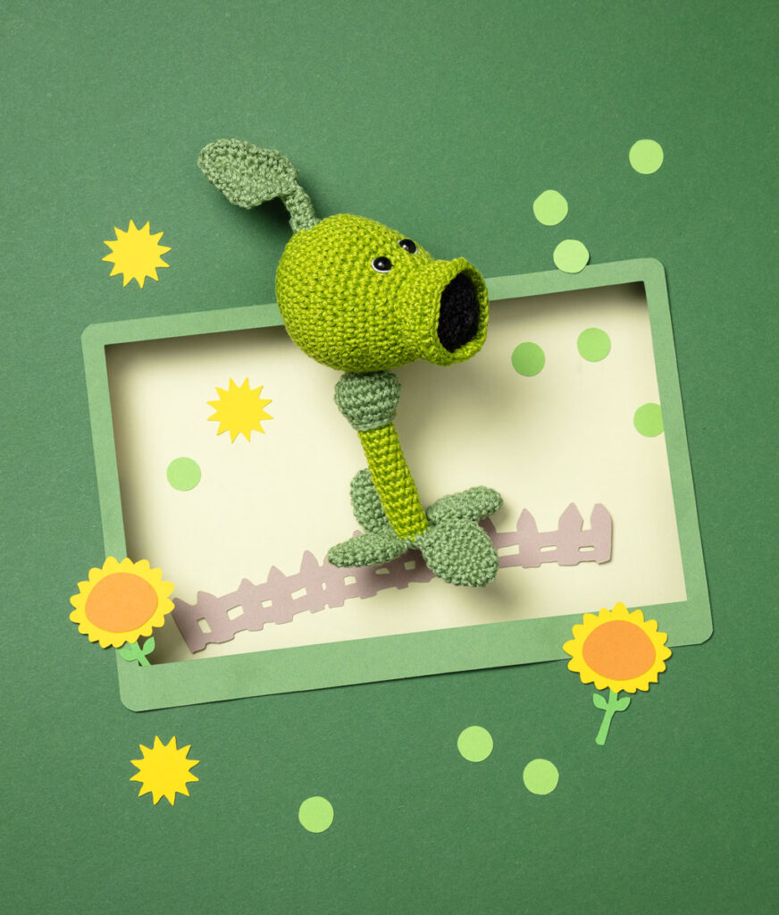 Produktfoto einer Amigurumi-Figur, die die Erbsenkanone aus dem Videospiel Plants vs. Zombies darstellt, arrangiert in einer Kulisse aus einem Papier Cut-Out in Form eines Bildschirms, dekoriert mit weiteren spieltypischen Paper-Art-Elementen
