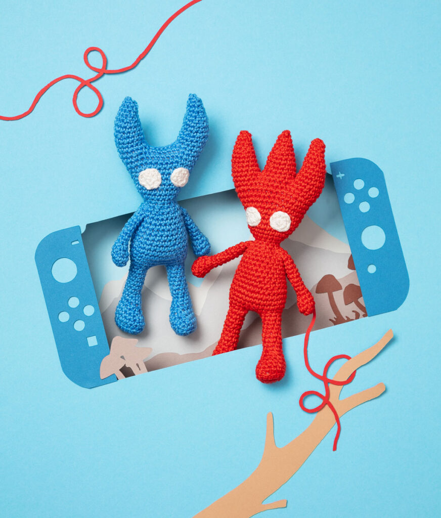 Produktfoto zweier Amigurumis, die die beiden Hauptfiguren aus dem Videospiel Unravel darstellt, arrangiert in einer Kulisse aus einem Papier Cut-Out in Form einer Switch, dekoriert mit weiteren spieltypischen Paper-Art-Elementen