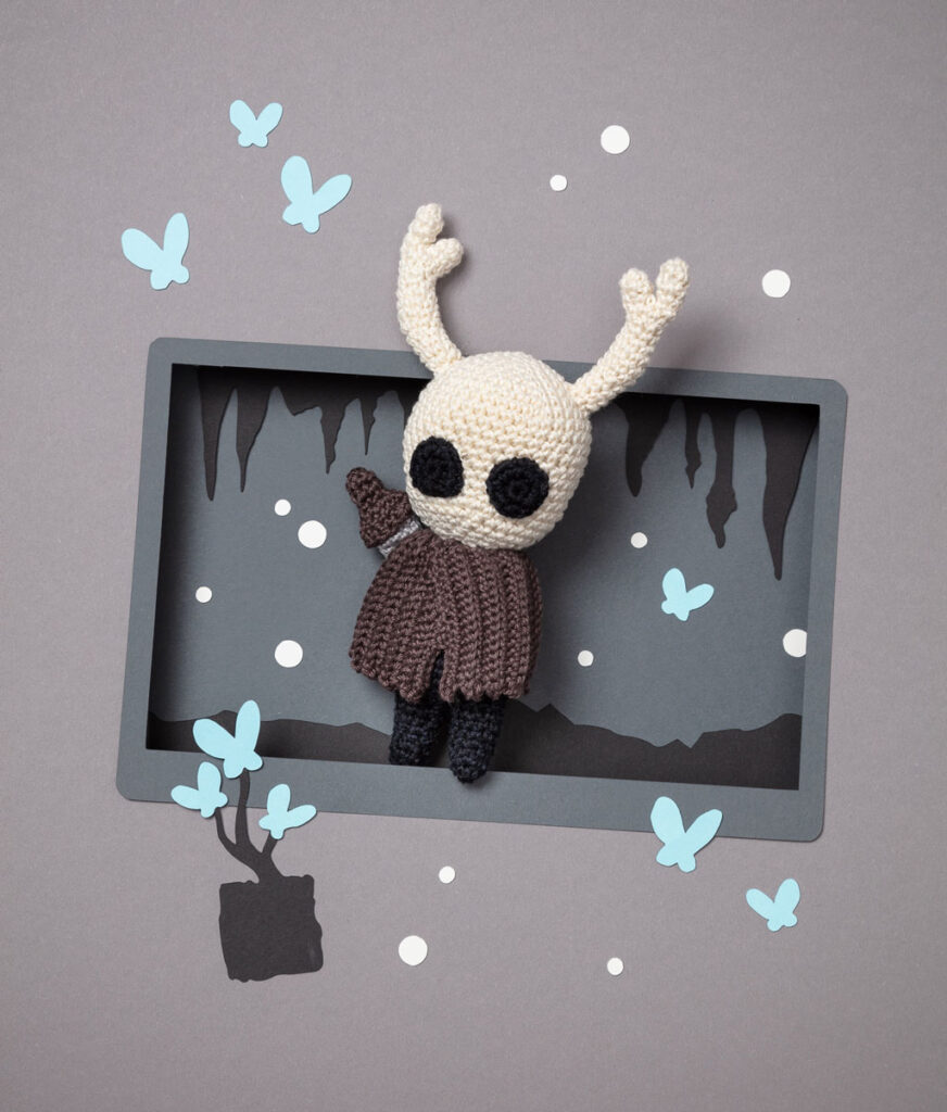 Produktfoto eines Amigurumis, das Hollow Knight aus dem Videospiel Hollow Knight darstellt, arrangiert in einer Kulisse aus einem Papier Cut-Out in Form eines Bildschirms, dekoriert mit weiteren spieltypischen Paper-Art-Elementen