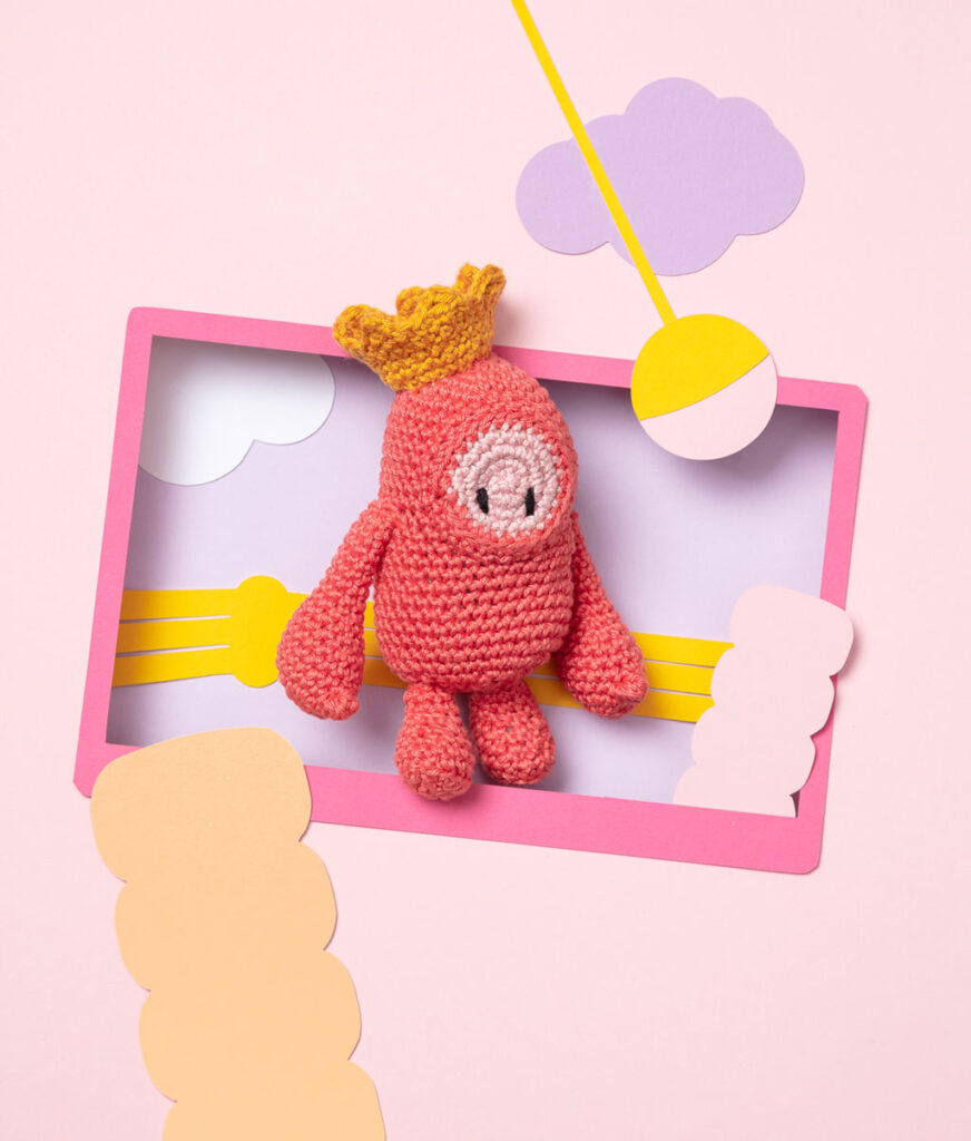 Produktfoto einer Amigurumi-Figur, die eine typische Figur aus dem Videospiel Fall Guys darstellt, arrangiert in einer Kulisse aus einem Papier Cut-Out in Form eines Bildschirms, dekoriert mit weiteren spieltypischen Paper-Art-Elementen