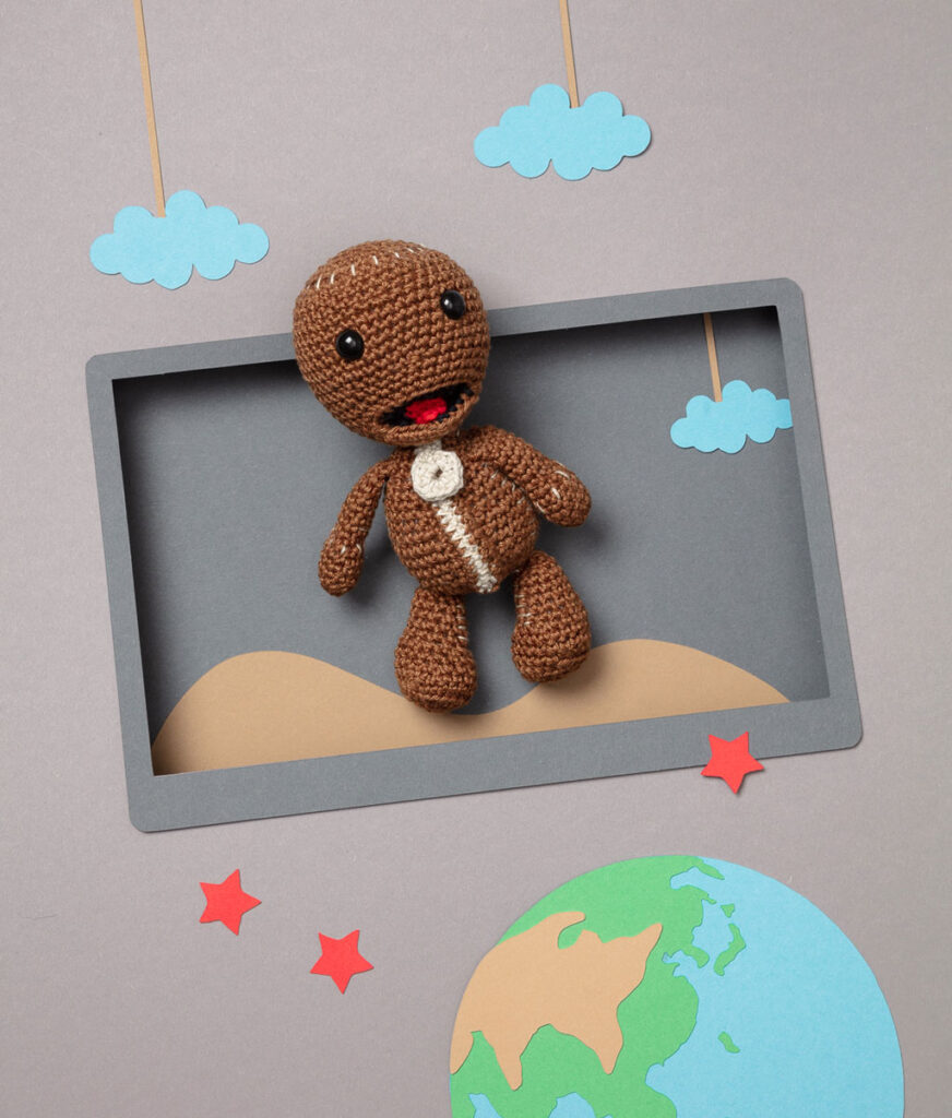 Produktfoto einer Amigurumi-Figur, die Sackboy aus dem Videospiel Little big planet darstellt, arrangiert in einer Kulisse aus einem Papier Cut-Out in Form eines Bildschirms, dekoriert mit weiteren spieltypischen Paper-Art-Elementen