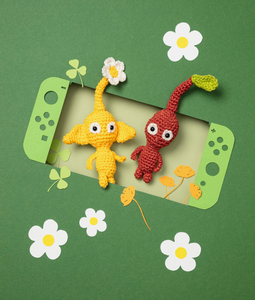 Produktfoto zweier Amigurumi-Figuren, die zwei Blattmännchen aus dem Videospiel Pikmin darstellen, arrangiert in einer Kulisse aus einem Papier Cut-Out in Form einer Switch, dekoriert mit weiteren spieltypischen Paper-Art-Elementen
