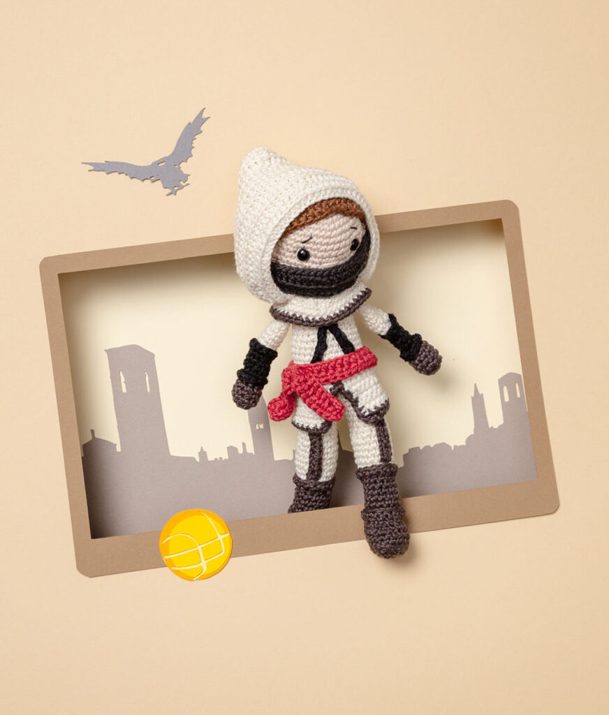 Produktfoto eines Amigurumis das Ezio Auditore da Firenze aus dem Videospiel Assassins Creed 2 darstellt, arrangiert in einer Kulisse aus einem Papier Cut-Out in Form eines Bildschirms, dekoriert mit weiteren spieltypischen Paper-Art-Elementen