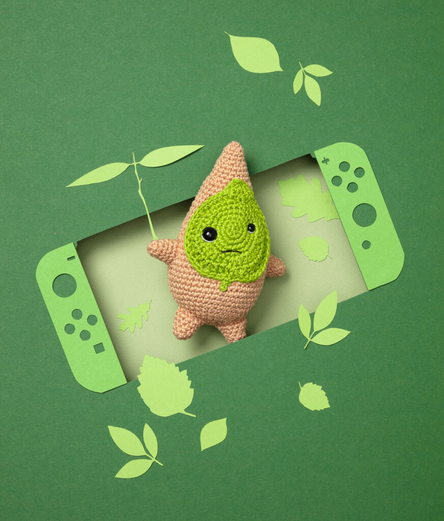 Produktfoto eines Amigurumis das Korok aus dem Videospiel Zelda darstellt, arrangiert in einer Kulisse aus einem Papier Cut-Out in Form einer Switch, dekoriert mit weiteren spieltypischen Paper-Art-Elementen