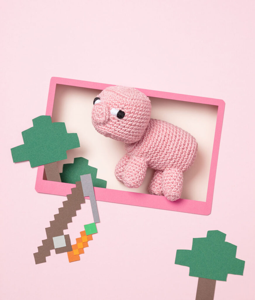 Produktfoto eines Amigurumis das ein Schwein aus dem Videospiel Minecraft darstellt, arrangiert in einer Kulisse aus einem Papier Cut-Out in Form eines Bildschirms, dekoriert mit weiteren spieltypischen Paper-Art-Elementen