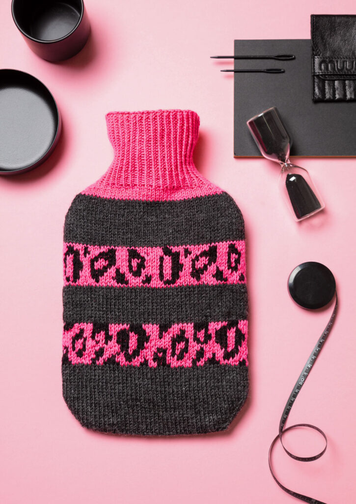 Produktfoto eines gestrickten Wärmflaschenbezugs mit pinkem Leo-Muster als Flatlay fotografiert mit schwarzen Deko-Elementen
